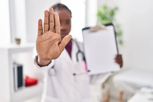 Beautiful black doctor woman holding clipboard with open hand doing stop sign with serious and confident expression, defense gesture