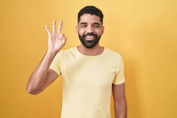自信と幸せ笑みを浮かべている間 髭を生やしたヒスパニック系の男は黄色の背景の上に立っていると指の数3を指して — ストック写真