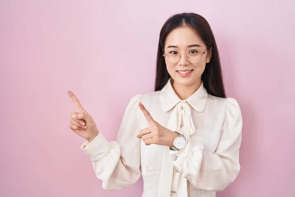 年轻的中国女人笑着站在粉色的背景上 看着摄像机 用双手和手指指向旁边 — 图库照片