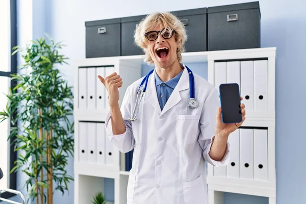 年轻的医生展示了智能手机屏幕 他把大拇指指向一边 笑着张开了嘴 — 图库照片