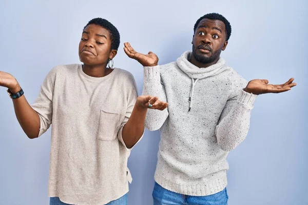 Stock image Young african american couple standing over blue background together clueless and confused expression with arms and hands raised. doubt concept. 