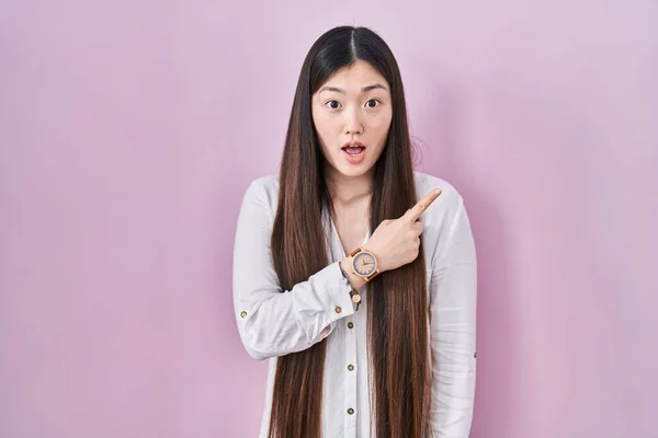中国の若い女性がピンクの背景に立って驚いて指で側面を指して 口を開けて驚いて表情 — ストック写真