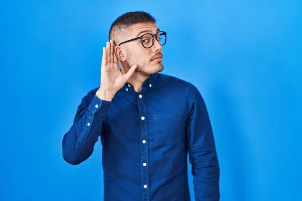 噂やゴシップに耳を傾ける耳の上に手で笑みを浮かべて青い背景に眼鏡をかけて若いヒスパニック系の男 聴覚障害の概念 — ストック写真