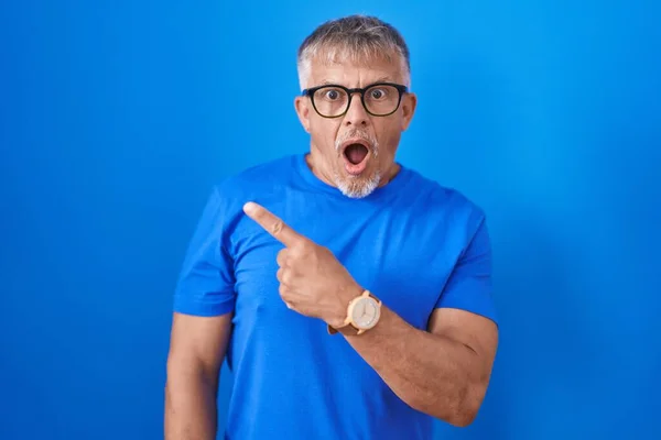 stock image Hispanic man with grey hair standing over blue background surprised pointing with finger to the side, open mouth amazed expression. 