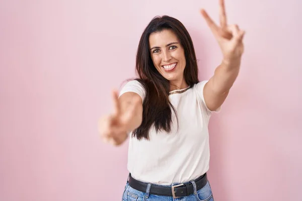 ピンクの背景に立っている若いブルネットの女性は 勝利のサインをして両手の指を見せて舌で笑っています — ストック写真