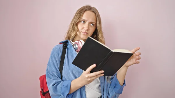 年轻的金发碧眼的女学生阅读书籍 对孤立的粉色背景表示怀疑 — 图库照片