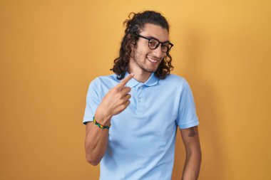 İspanyol asıllı genç adam sarı arka planda duruyor. Buraya el kol hareketi yaparak mutlu bir şekilde karşılamaya ve gülümsemeye davet ediyor. 