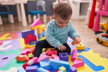 Adorable hispanic baby playing with construction blocks sitting on floor at kindergarten