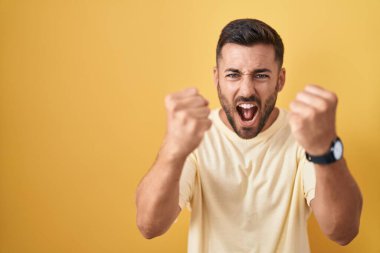 Sarı arka planda dikilen yakışıklı İspanyol adam öfkeyle bağırırken kızgın ve kızgın yumruklar sallıyor. öfke ve agresif konsept. 