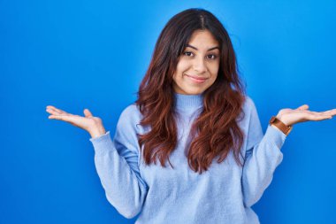 Mavi arka planda dikilen Latin kökenli genç kadın elleri ve kolları havada, kafası karışmış bir ifade takınıyor. şüphe kavramı. 