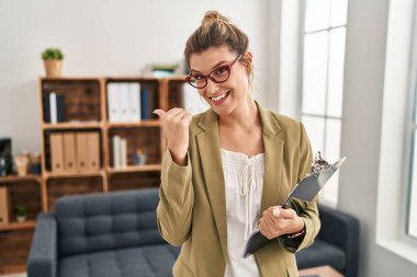 Young woman working at consultation office pointing to the back behind with hand and thumbs up, smiling confident 