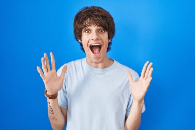 Hispanic young man standing over blue background celebrating crazy and amazed for success with arms raised and open eyes screaming excited. winner concept 