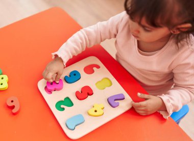 Adorable hispanic girl playing with maths puzzle game sitting on table at kindergarten