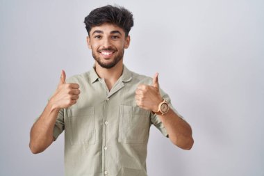 Beyaz arka plan başarı tabelasının üzerinde duran sakallı bir Arap. Elleriyle olumlu bir hareket yapıyor. Gülümsüyor ve mutlu. Neşeli ifade ve kazanan jest. 