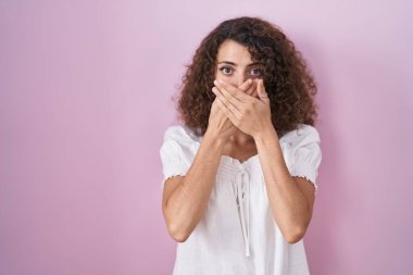 Pembe arka planda duran kıvırcık saçlı İspanyol kadın yanlışlıkla elleri ile ağzını kapatırken şok oldu. gizli kavram. 
