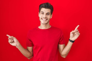 Young hispanic man standing over red background smiling confident pointing with fingers to different directions. copy space for advertisement 