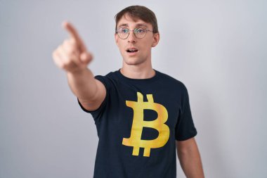 Bitcoin tişörtlü, beyaz tenli, sarışın bir adam. Parmakları önde, ağzı açık, şaşırmış, ön tarafta bir şey var. 