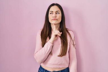 Young brunette woman standing over pink background thinking concentrated about doubt with finger on chin and looking up wondering 