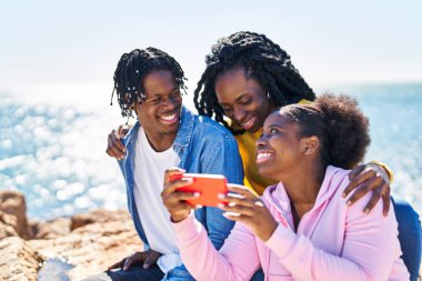 African american friends using smartphone sitting on rock at seaside
