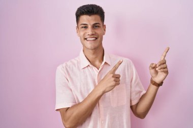 Young hispanic man standing over pink background smiling and looking at the camera pointing with two hands and fingers to the side. 