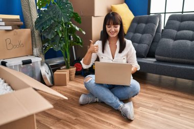 Genç esmer kadın yeni evinde dizüstü bilgisayarla oturmuş gülümsüyor, mutlu ve pozitif gülümsüyor, başparmağıyla mükemmel ve onay işareti yapıyor. 