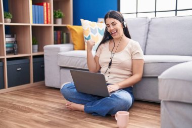 Young hispanic woman using laptop at home pointing thumb up to the side smiling happy with open mouth 