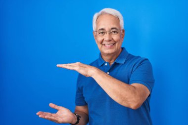 Orta yaşlı, gri saçlı, mavi arka planda duran, elleri büyük ve büyük boy işareti gösteren, ölçü sembolü olan bir adam. Kameraya bakıp gülümsüyor. ölçüm kavramı. 