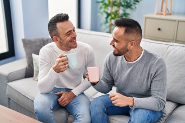Two men couple drinking coffee sitting on sofa at home