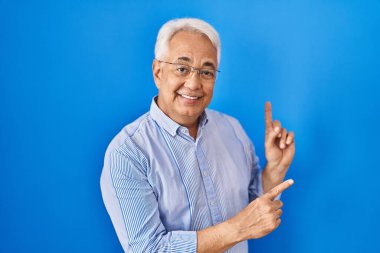 Hispanic senior man wearing glasses smiling and looking at the camera pointing with two hands and fingers to the side. 