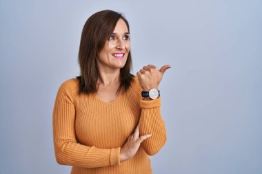 Orta yaşlı esmer kadın turuncu kazak giyiyor mutlu yüzüyle gülümsüyor ve başparmağıyla yan tarafı işaret ediyor.. 