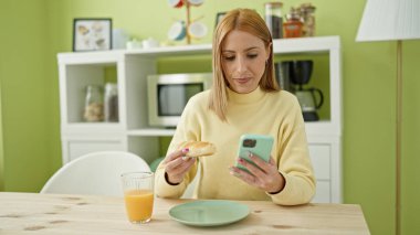 Young blonde woman using smartphone having breakfast at home