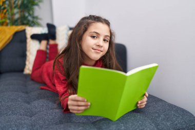 Adorable hispanic girl reading book lying on sofa at home