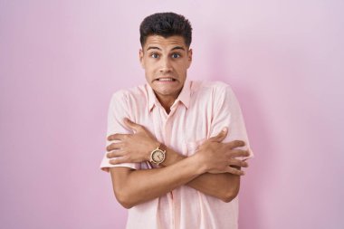 Young hispanic man standing over pink background shaking and freezing for winter cold with sad and shock expression on face 