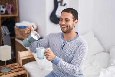 Young man drinking cup of coffee sitting on bed at bedroom