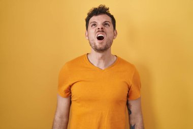 Young hispanic man standing over yellow background angry and mad screaming frustrated and furious, shouting with anger. rage and aggressive concept. 