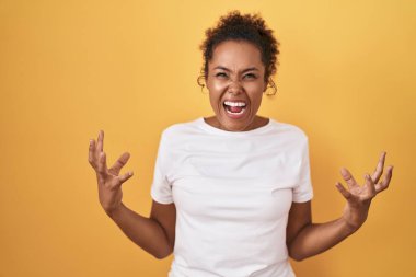 Young hispanic woman with curly hair standing over yellow background crazy and mad shouting and yelling with aggressive expression and arms raised. frustration concept. 