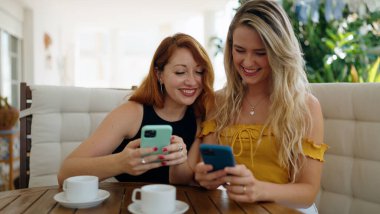Two women using smartphones and drinking coffee sitting on table at home terrace