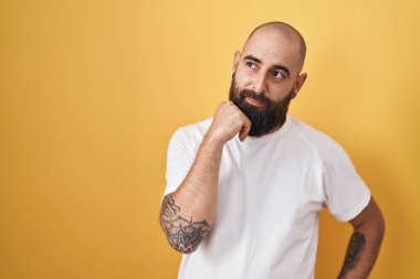 Sakallı ve dövmeli genç İspanyol bir adam. Sarı arka planda duruyor. Elleri çenesinde soru sormayı düşünüyor. Düşünceli bir ifade. Düşünceli bir yüzle gülümsüyor. şüphe kavramı. 