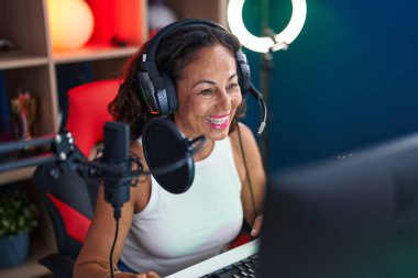 Middle age woman streamer smiling confident sitting on table at gaming room