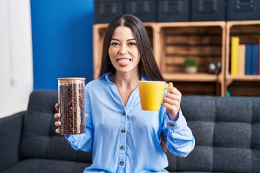 Genç esmer kadın elinde kahve çekirdekleri ve kahve fincanı tutarken komik bir ifadeyle dilini dışarı çıkartıyor.. 