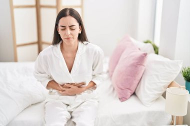 Young hispanic woman suffering for menstrual pain sitting on bed at bedroom