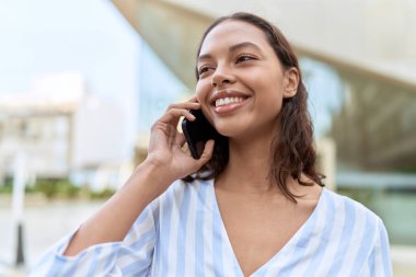 Genç Afrikalı Amerikalı kadın gülümsüyor. Kendine güveni tam. Sokakta akıllı telefondan konuşuyor.