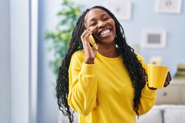 Afro-Amerikalı kadın kahve içiyor ve evde akıllı telefondan konuşuyor.