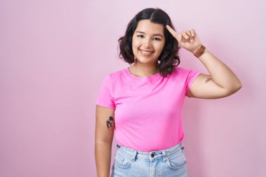 Young hispanic woman standing over pink background smiling pointing to head with one finger, great idea or thought, good memory 