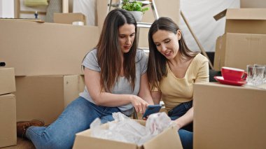 İki kadın yeni evinde yerde otururken akıllı telefon kullanıyor.