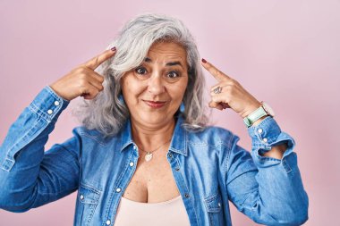 Pembe arka planda duran gri saçlı orta yaşlı kadın iki eliyle başını gösteren gülümseme, harika bir fikir ya da düşünce, iyi bir hafıza 