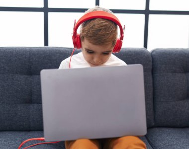 Adorable hispanic boy using laptop and headphones sitting on sofa at home