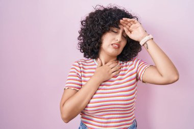 Orta Doğulu genç bir kadın pembe arka planda duruyor. Hastalık, ateş, grip ve virüs için alnına dokunuyor. 