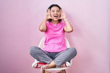 Hispanic young woman sitting on chair over pink background smiling cheerful playing peek a boo with hands showing face. surprised and exited 