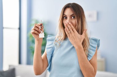 Young hispanic woman holding keys of new home covering mouth with hand, shocked and afraid for mistake. surprised expression 